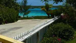 A coastal road with a white fence, lush greenery, and a view of turquoise waters and a distant island. - Ranked by VRBO/HomeAway Top 10 Most Unique Rentals Worldwide, Walk to Town. (Hatchet Bay, Eleuthera, Bahamas)