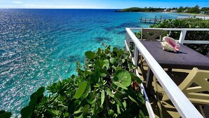 A clear view of a turquoise ocean from a wooden deck with a white railing, overlooking a small island with buildings and a dock. - Ranked by VRBO/HomeAway Top 10 Most Unique Rentals Worldwide, Walk to Town. (Hatchet Bay, Eleuthera, Bahamas)