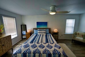 Une chambre à coucher comprenant un lit, un ventilateur de plafond, une table de chevet, une commode et une fenêtre avec des stores.