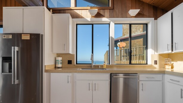 A modern kitchen with white cabinets, stainless steel appliances, a large window with a view of the sea, and a balcony with a railing. - 7BR Dillon Beach Oceanfront Home - Hot Tub + Game Room (Dillon Beach)