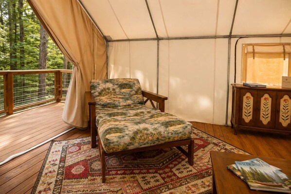 A tented room with a bed, a wooden bench, a table with books, and a view of trees.