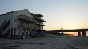 Een meerhoog gebouw aan het strand, met een trap die naar het strand leidt, een brug op de achtergrond en een zonsondergang.