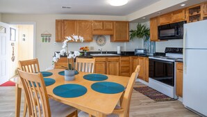 Een moderne keuken met houten kastjes, een magnetron, fornuis en een koelkast. Er is een eettafel met stoelen en een vaas met bloemen.