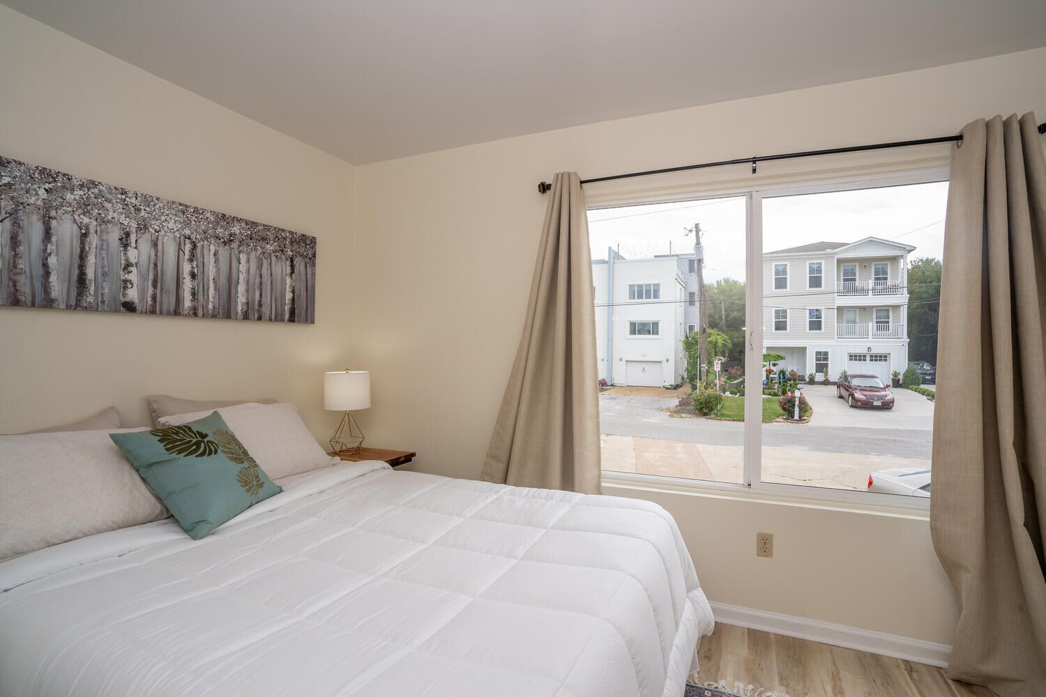 A bedroom with a large bed, a nightstand with a lamp, a window with curtains, and a view of a building outside.