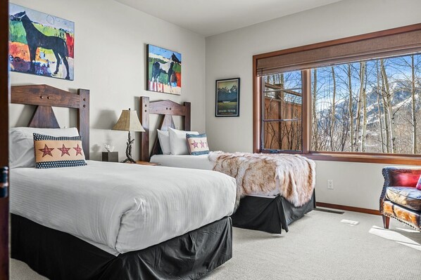 A bedroom with two beds, a large window with a view of trees and mountains, and artwork on the walls.