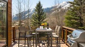 Una terraza de madera con una mesa, sillas, una parrilla y vistas a montañas nevadas.