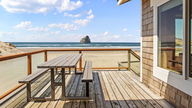 A wooden deck with picnic tables overlooking a beach with a rock formation in the distance.
