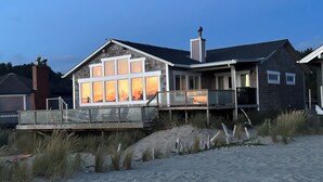 A modern house with large windows, a balcony, and a sandy beach in the foreground.