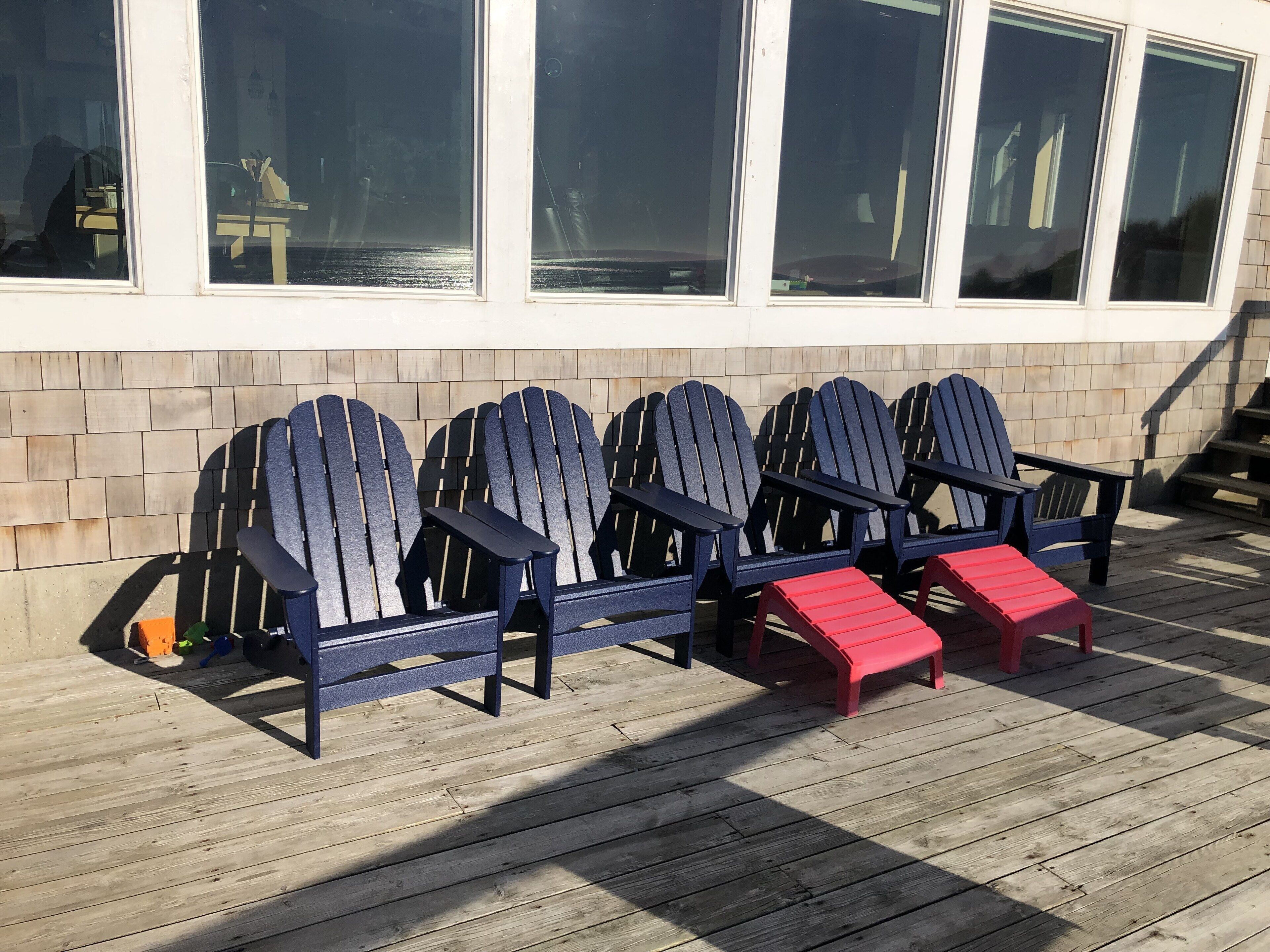 A row of Adirondack chairs on a wooden deck with a brick wall and windows in the background.