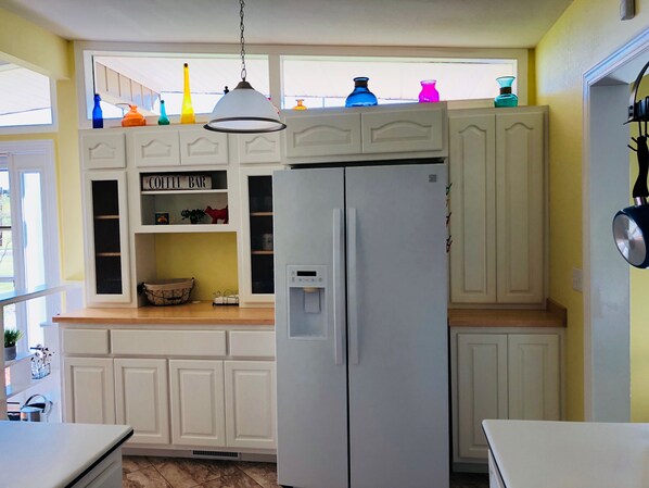 Una cocina con gabinetes blancos, un refrigerador blanco, un letrero que dice ‘cafetería’ y jarrones coloridos en el alféizar de la ventana.