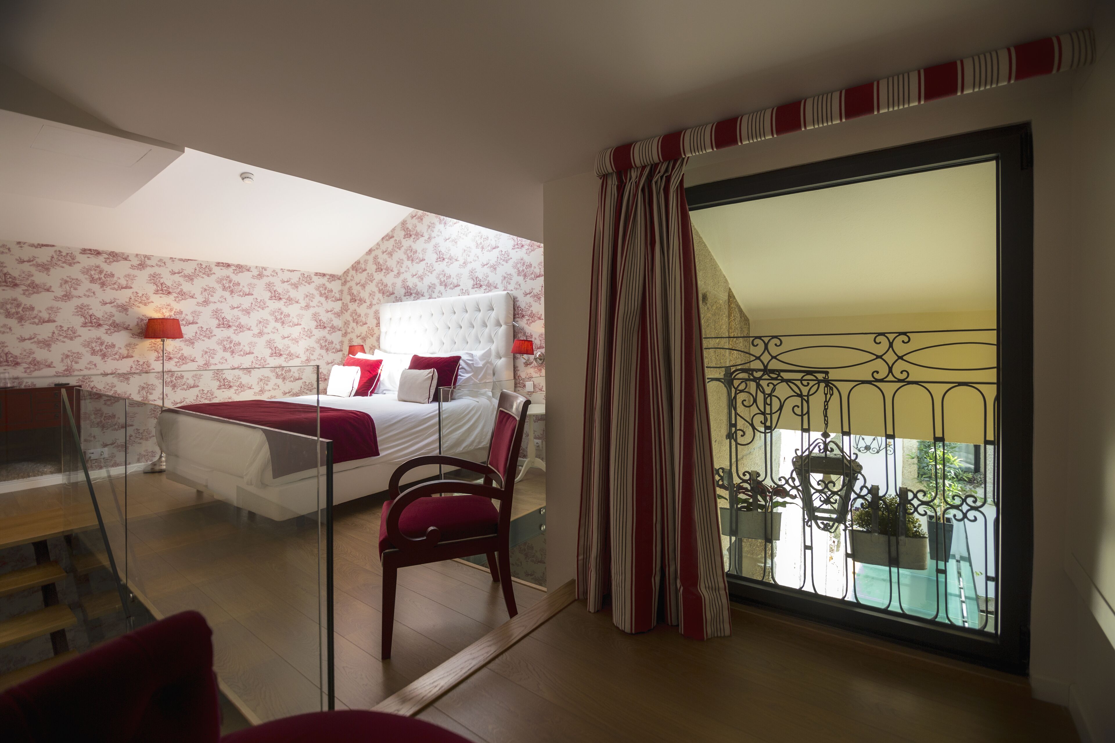 A bedroom with a bed, a chair, a window with curtains, and a balcony with plants.