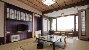 Una habitación japonesa tradicional con suelo de tatami, televisor, mesa y vistas a través de amplios ventanales.