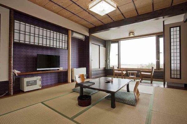 A traditional Japanese room with tatami flooring, a television, a table, and a view through large windows.