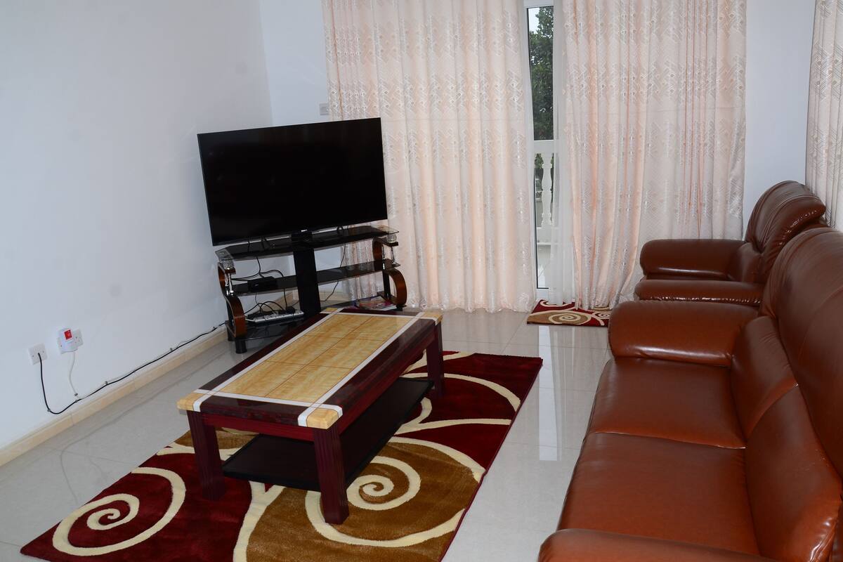 A living room with a TV, a wooden coffee table, a brown leather sofa, and patterned curtains.