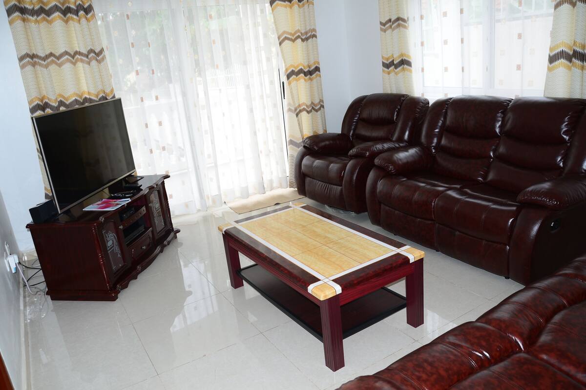A living room with a leather sofa, a wooden coffee table, a TV on a wooden stand, and a window with patterned curtains.