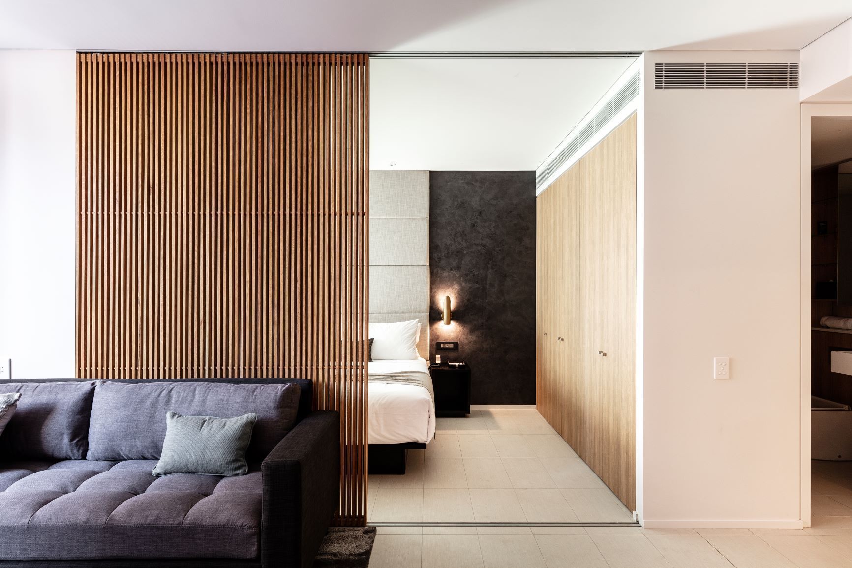 A modern hotel room with a sofa, a bed, a nightstand, and a wooden partition.