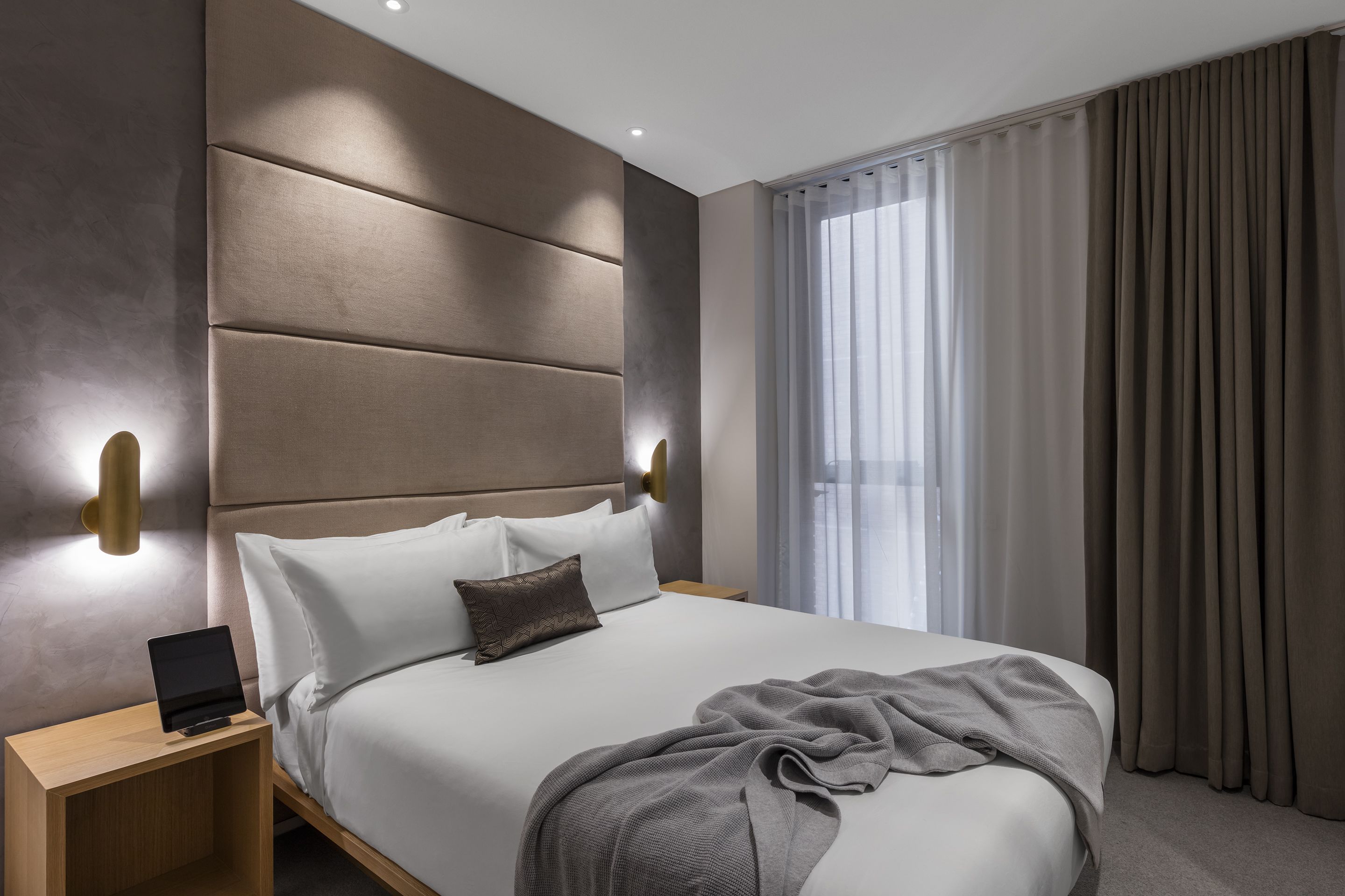 Una habitación de hotel moderna con una cama grande, una mesita de noche con lámpara y vistas al exterior a través de cortinas translúcidas.