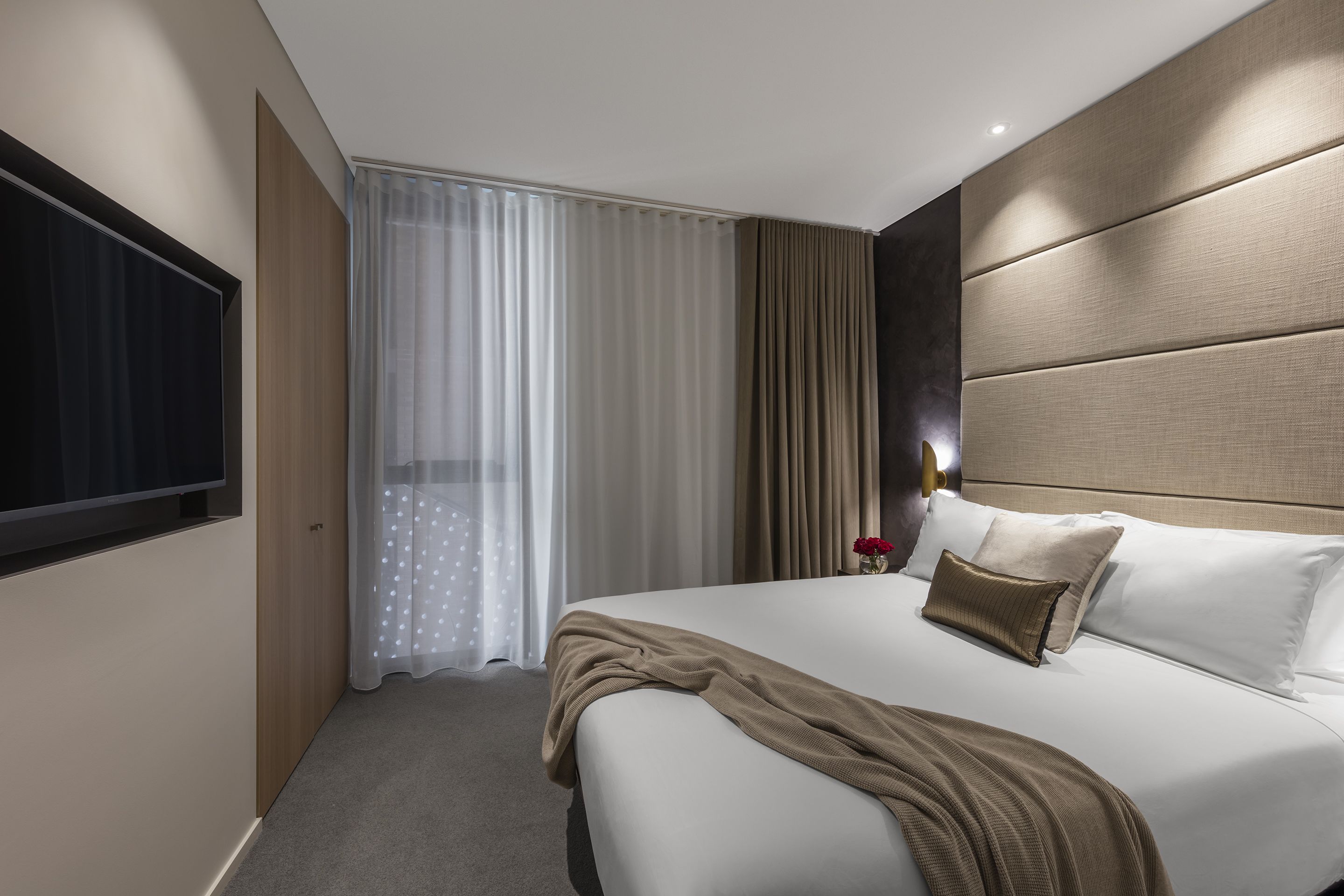Et moderne hotelværelse med en stor seng, et fladskærms-tv ophængt på væggen og et vindue med lette gardiner.