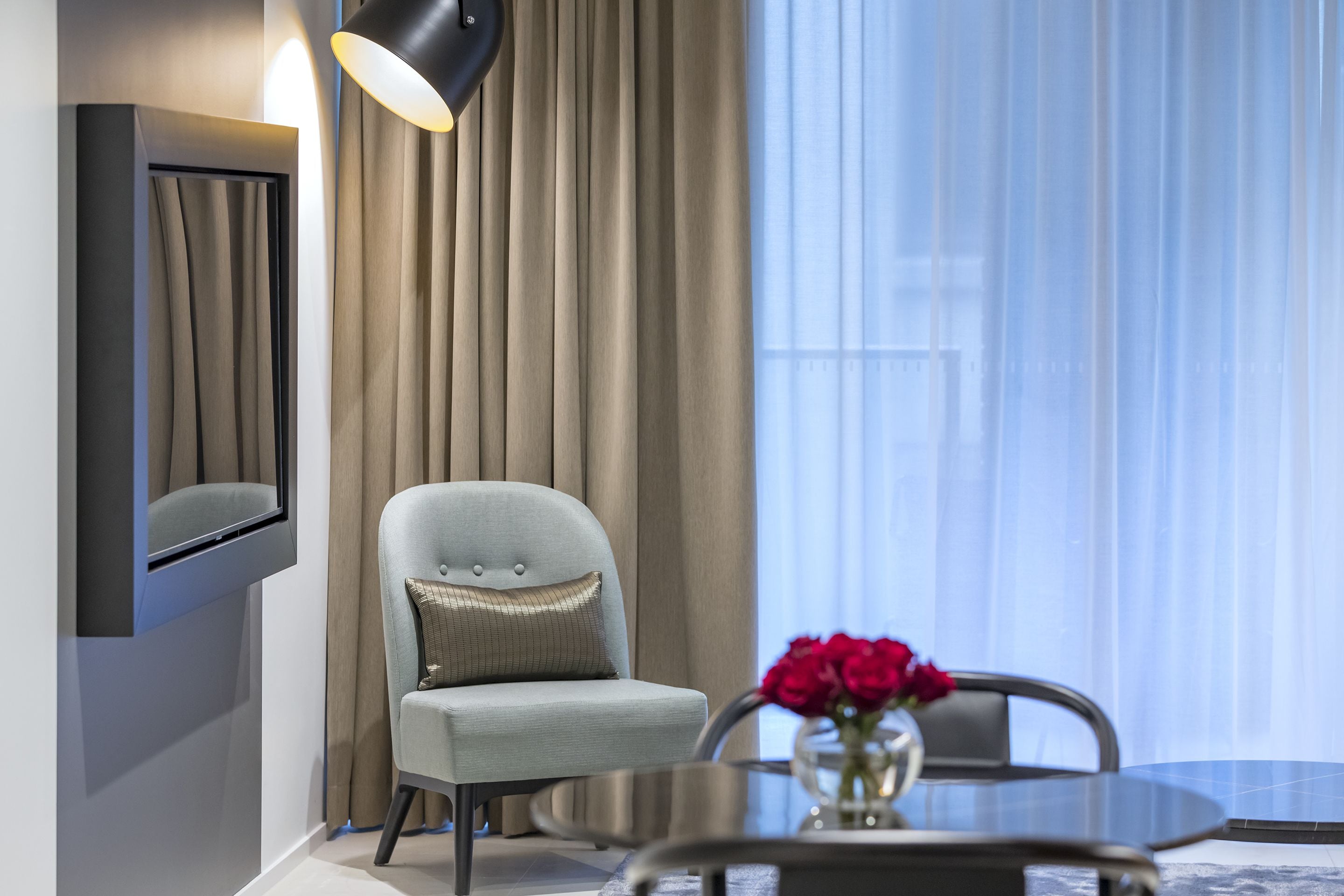 Una camera moderna con una TV a schermo piatto montata a parete, una poltrona grigio chiaro con un cuscino, un tavolino rotondo in vetro con un vaso di fiori rossi e un'ampia finestra con tende leggere.