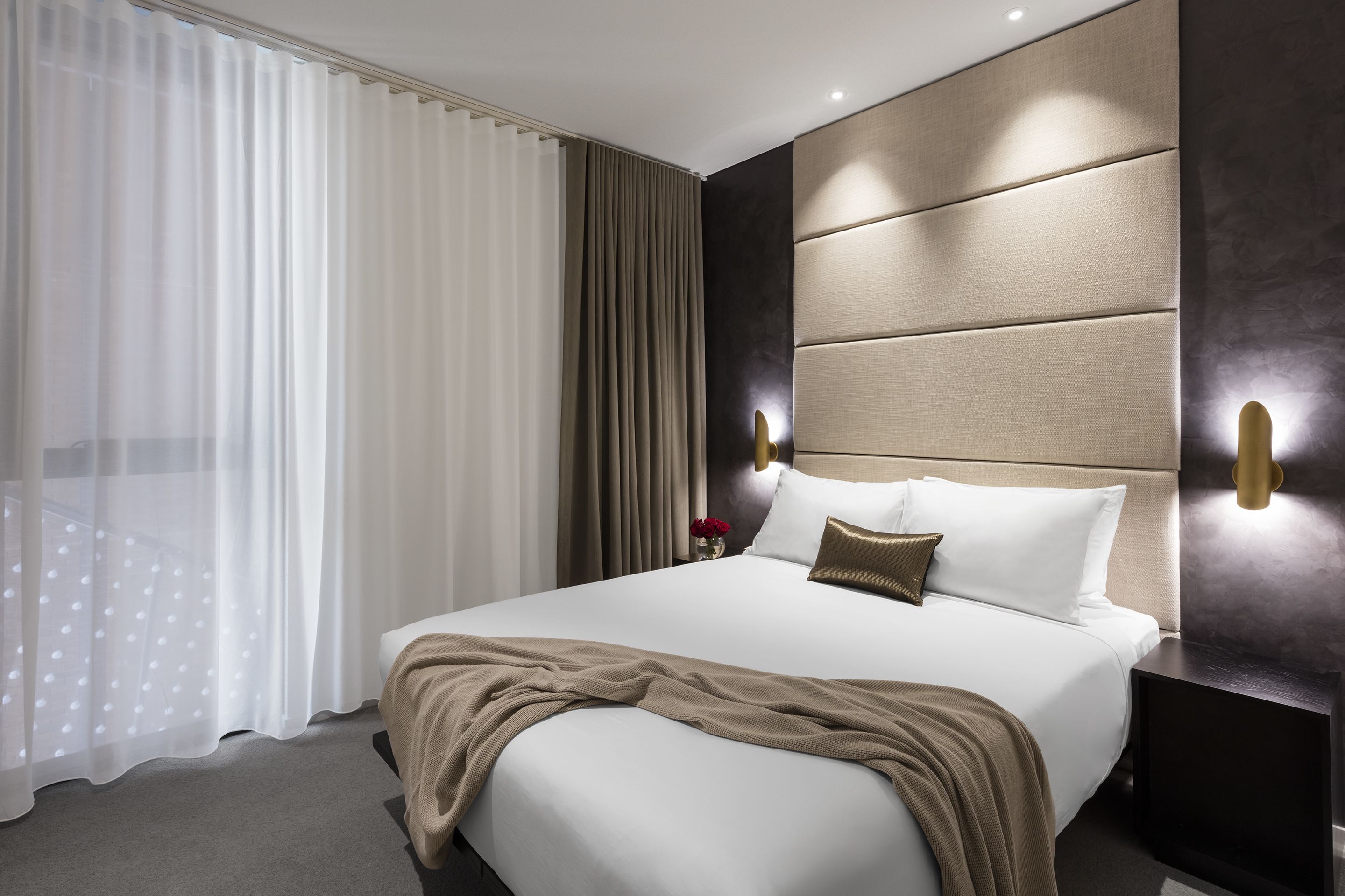 A modern hotel room with a large bed, a headboard with built-in lighting, a bedside table, and a view of the city through sheer curtains.