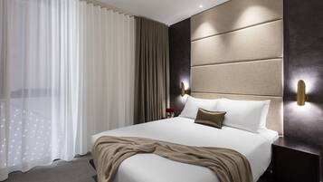 A modern hotel room with a large bed, a headboard with built-in lighting, a bedside table, and a view of the city through sheer curtains.