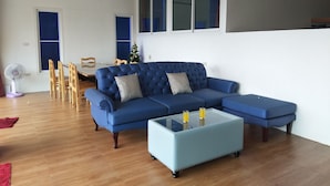 A living room with a blue sectional sofa, a glass coffee table, wooden chairs, and a small table with two glasses. - White House Mountian Beach Resort (Klaeng)