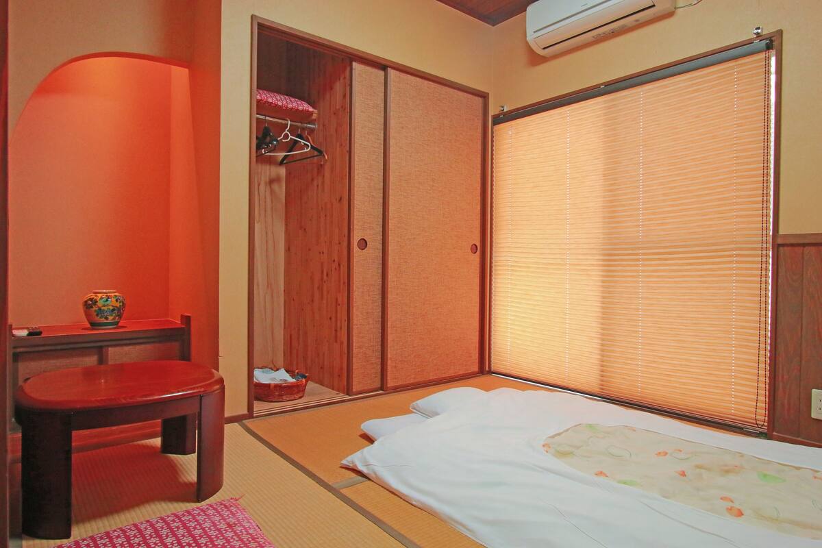 A traditional Japanese room with tatami flooring, a futon bed, a wooden wardrobe, and a sliding door with blinds.