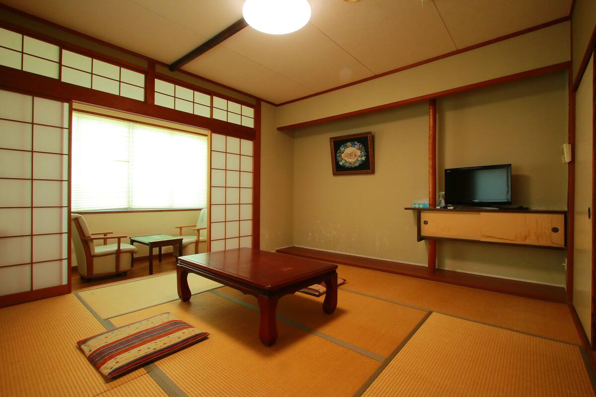 A traditional Japanese room with tatami flooring, a low wooden table, a television on a shelf, and a shoji screen.