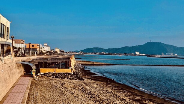 Une zone côtière dotée d’une promenade pavée, d’une plage rocheuse et offrant une vue sur la mer et les collines lointaines.