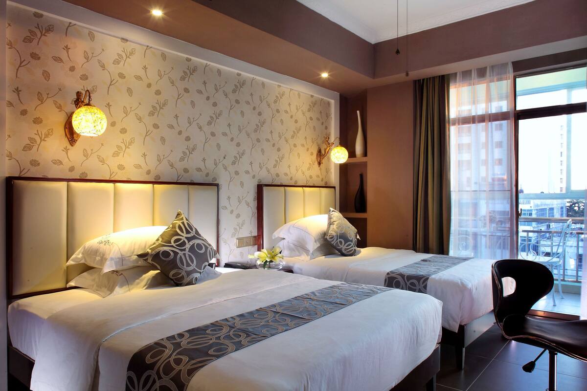 A hotel room with two beds, a chair, a large window with a view, and decorative wall sconces.
