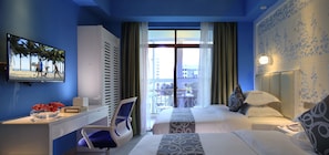A hotel room with a bed, a desk with a chair, a TV displaying a beach scene, and a balcony with a view of the city. - Care Hotel Coast Collection (Sanya)