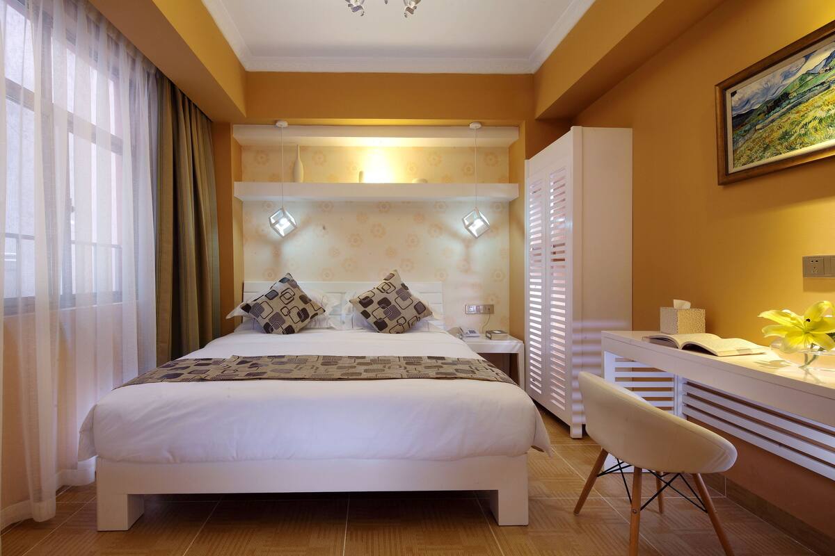 A neatly arranged hotel room with a large bed, a bedside table with a lamp, a desk with a chair, and a painting on the wall.