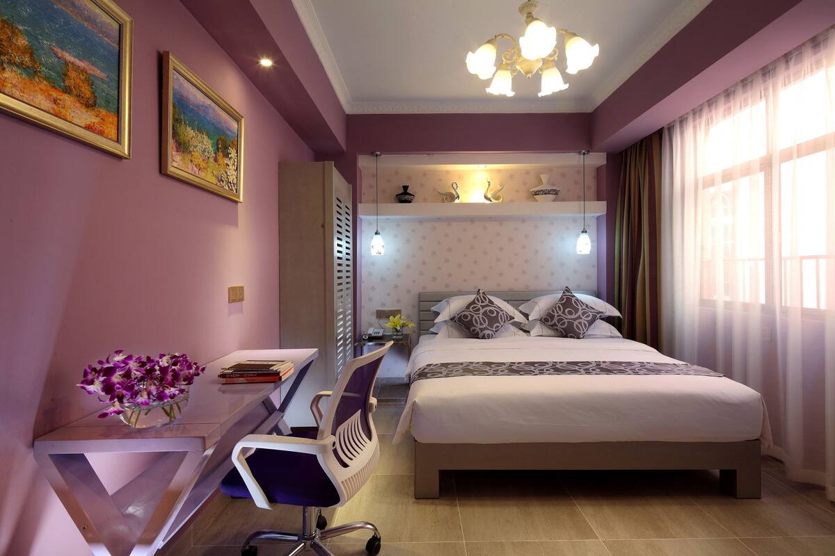 A bedroom with a bed, desk, chair, and a small table with a vase of flowers.