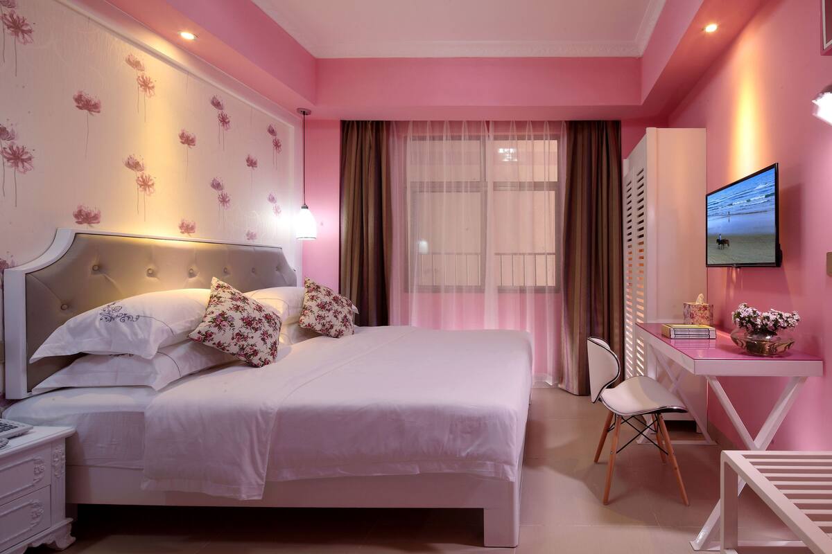 A bedroom with a bed, bedside table, lamp, desk, chair, and a TV on the wall.