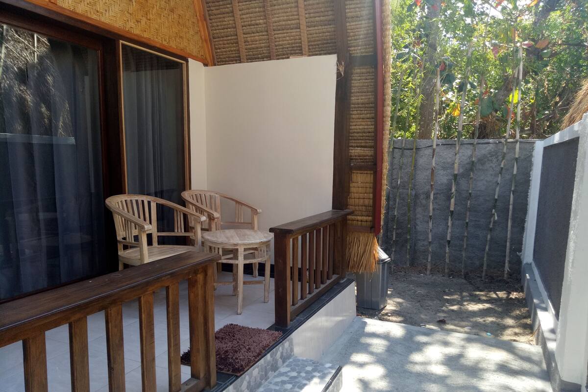 Un área de estar al aire libre techada, con sillas de madera y una mesa, una alfombra pequeña y una valla de bambú.