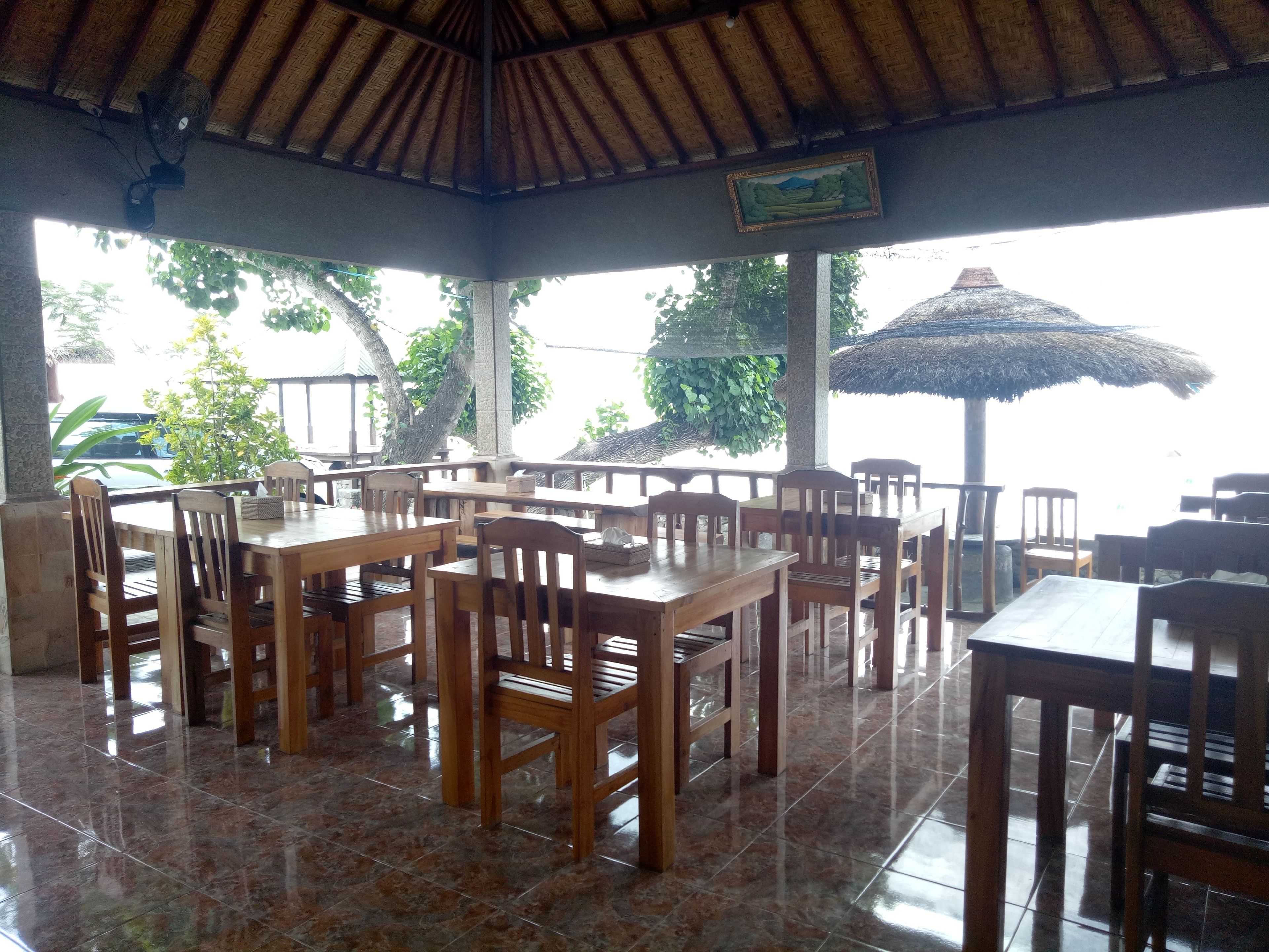 A covered outdoor dining area with wooden tables and chairs, a thatched roof, and a view of the sea.