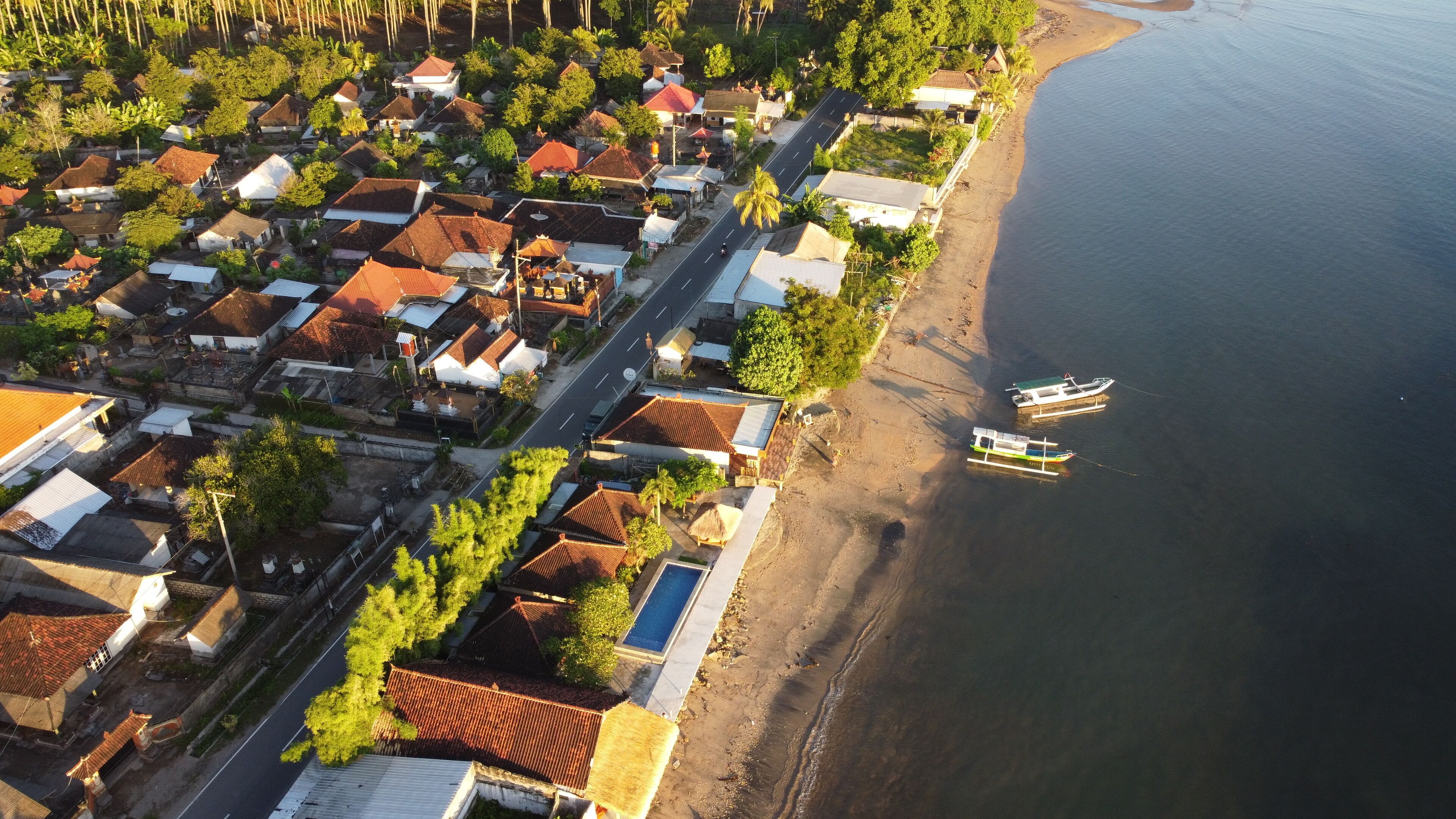An aerial view of a coastal village with traditional houses, a main road, and boats docked near the shore.