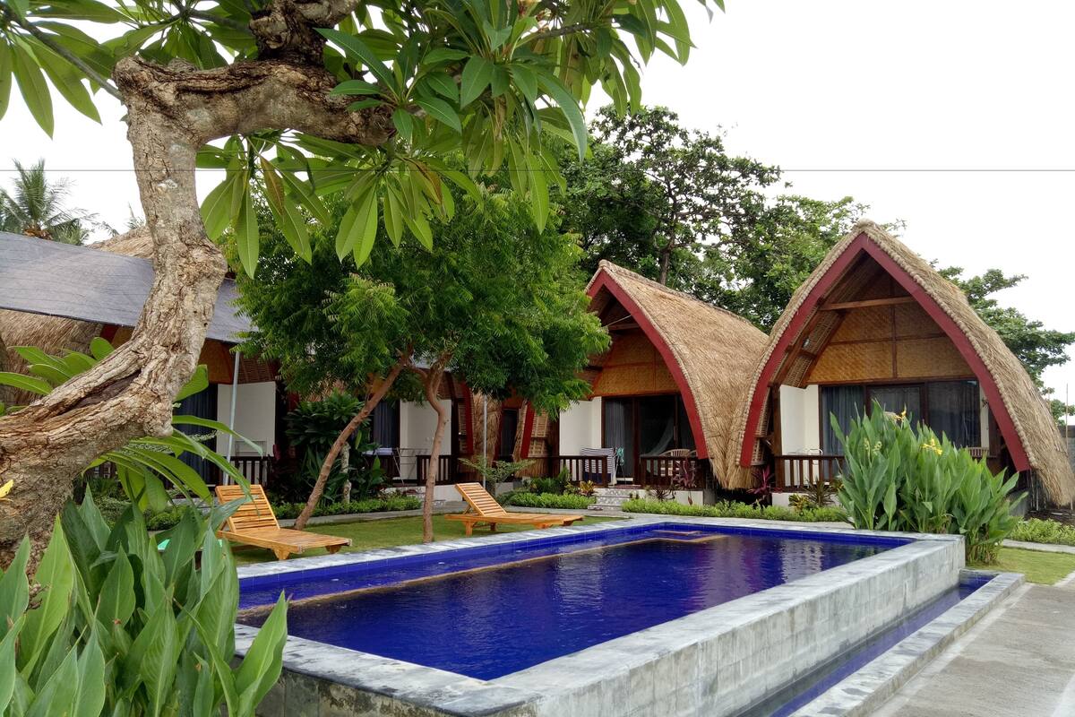 Un área de piscina con piscina azul, sillas de descanso y cabañas con techo de paja.