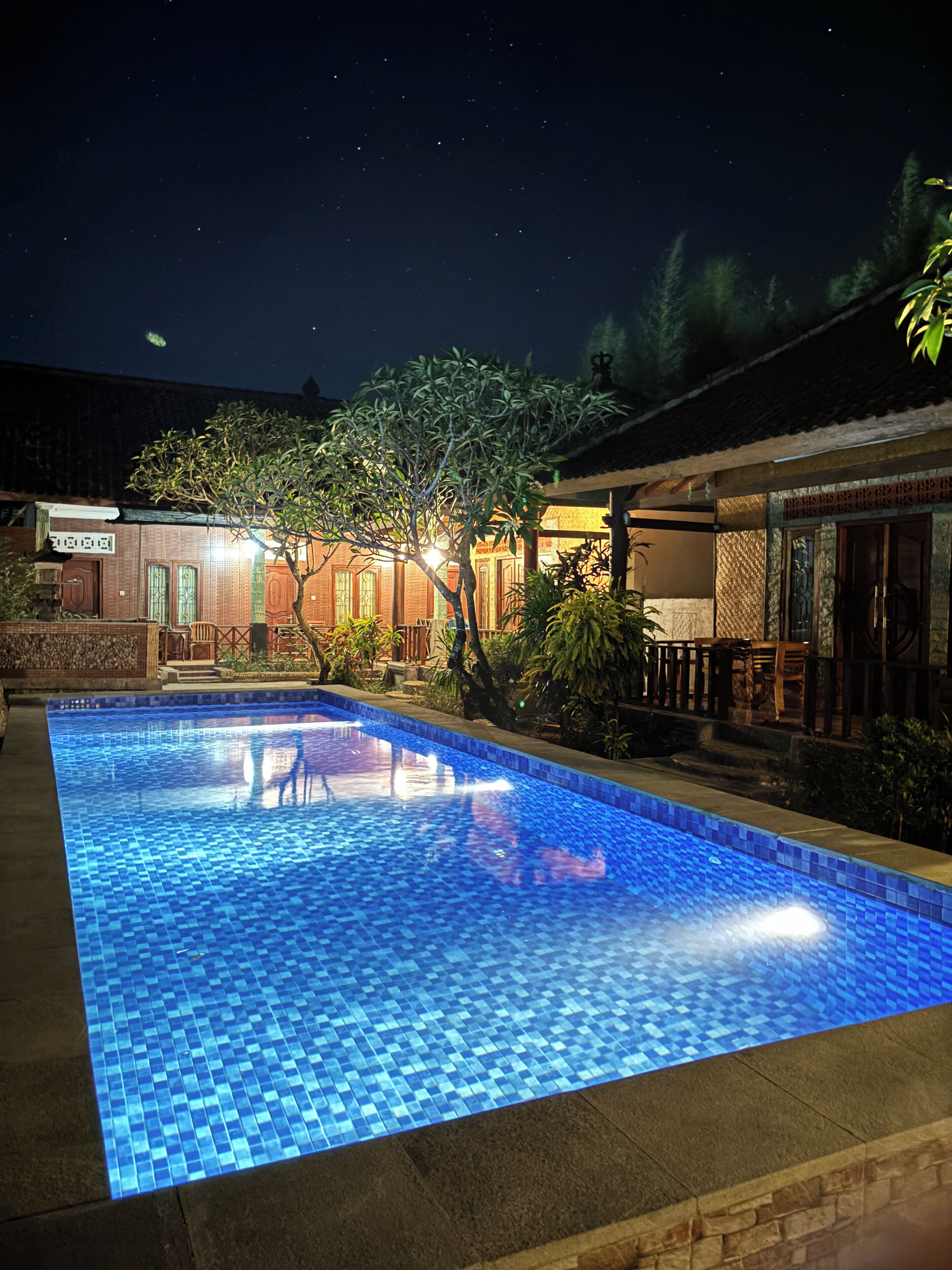 A nighttime swimming pool with a tiled surface, surrounded by a building with traditional architecture and a small garden area.
