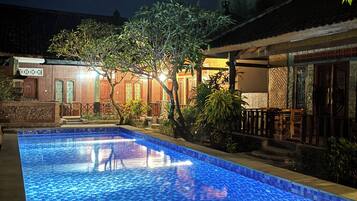 A nighttime swimming pool with a tiled surface, surrounded by a building with traditional architecture and a small garden area.