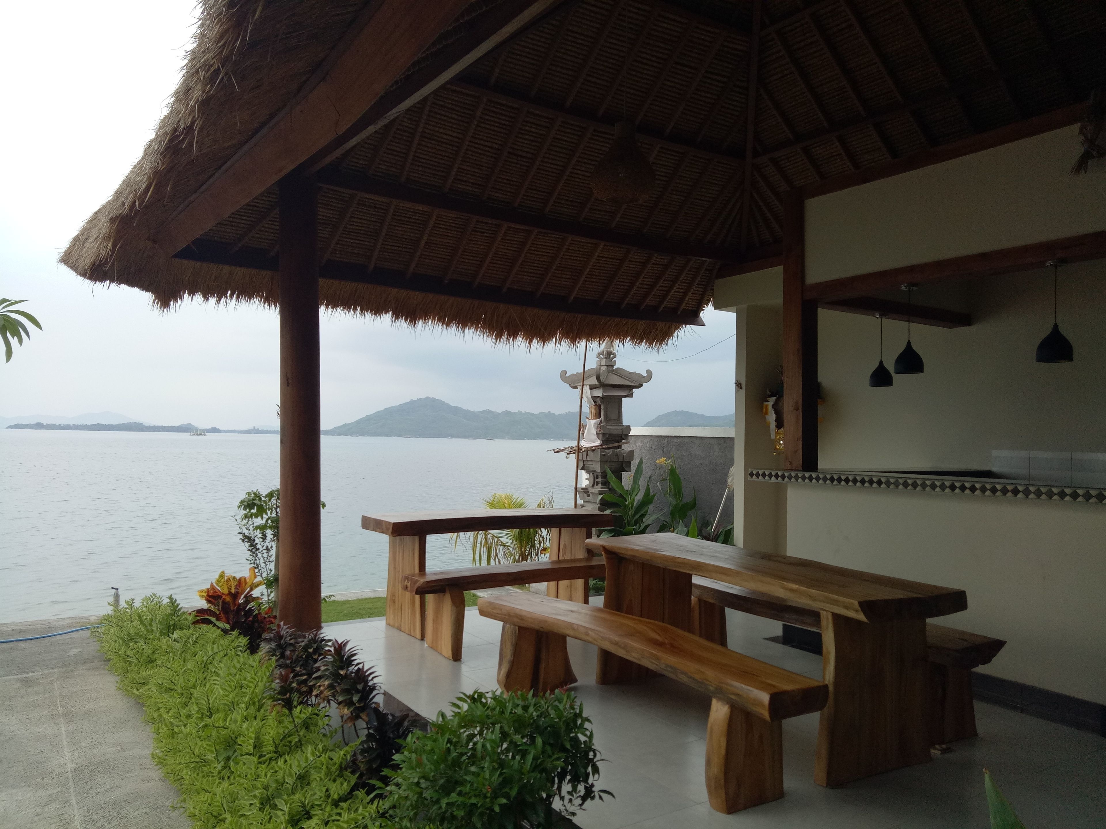 A wooden bench and table under a thatched roof, overlooking a calm sea with a distant mountain.