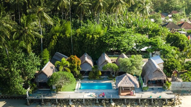 Vue aérienne d’un complexe hôtelier avec des bungalows aux toits de chaume, une piscine et une plage.