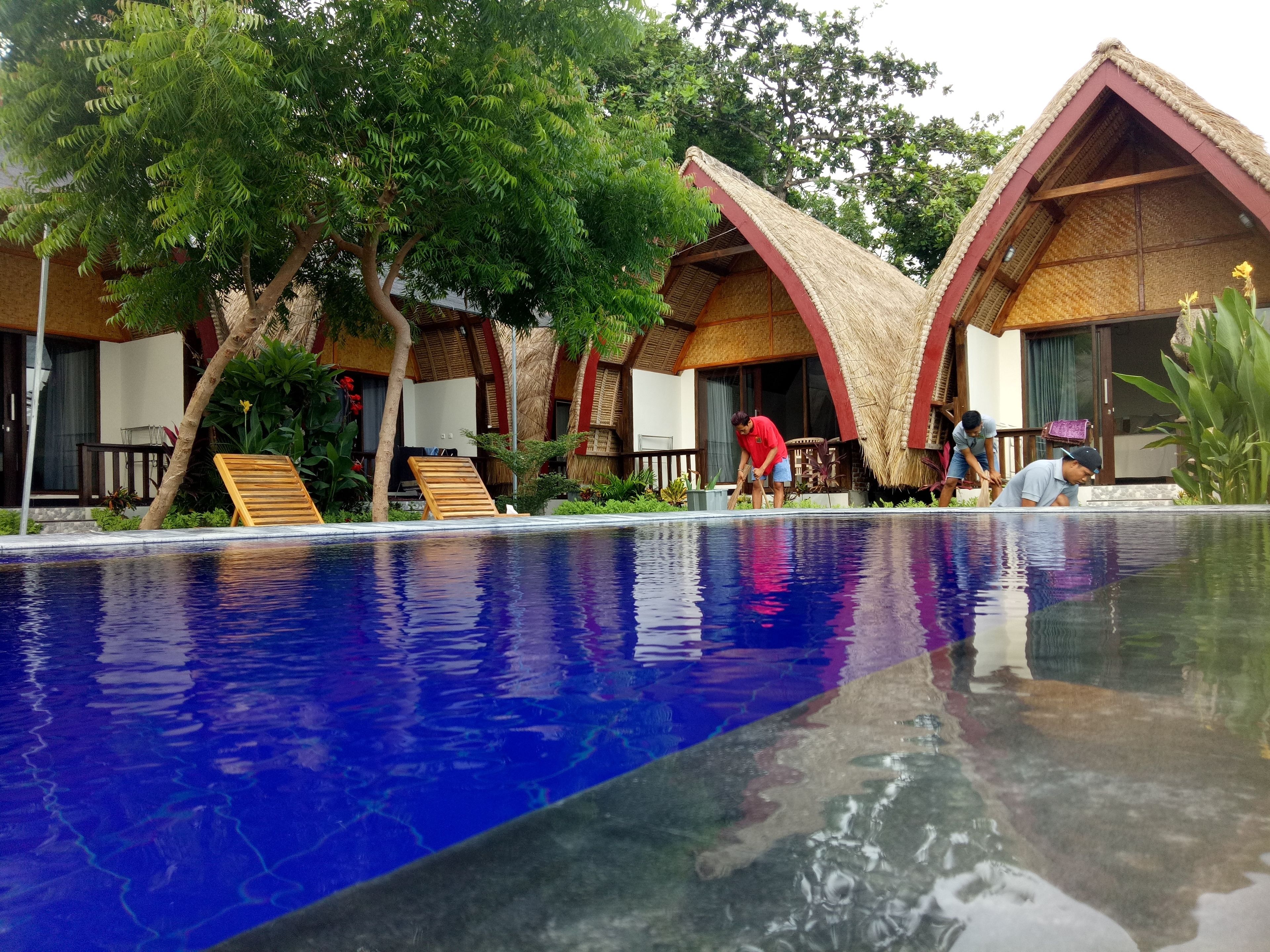 Un espace aménagé au bord d’une piscine à l’eau d’un bleu limpide, avec des chaises longues en bois et des bâtiments aux toits de chaume.