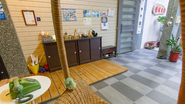 A reception area with a checkered floor, a wicker sofa, a wooden table with a green telephone, a bulletin board with various notices, and a brown cabinet. - DingDing Home (Hengchun)