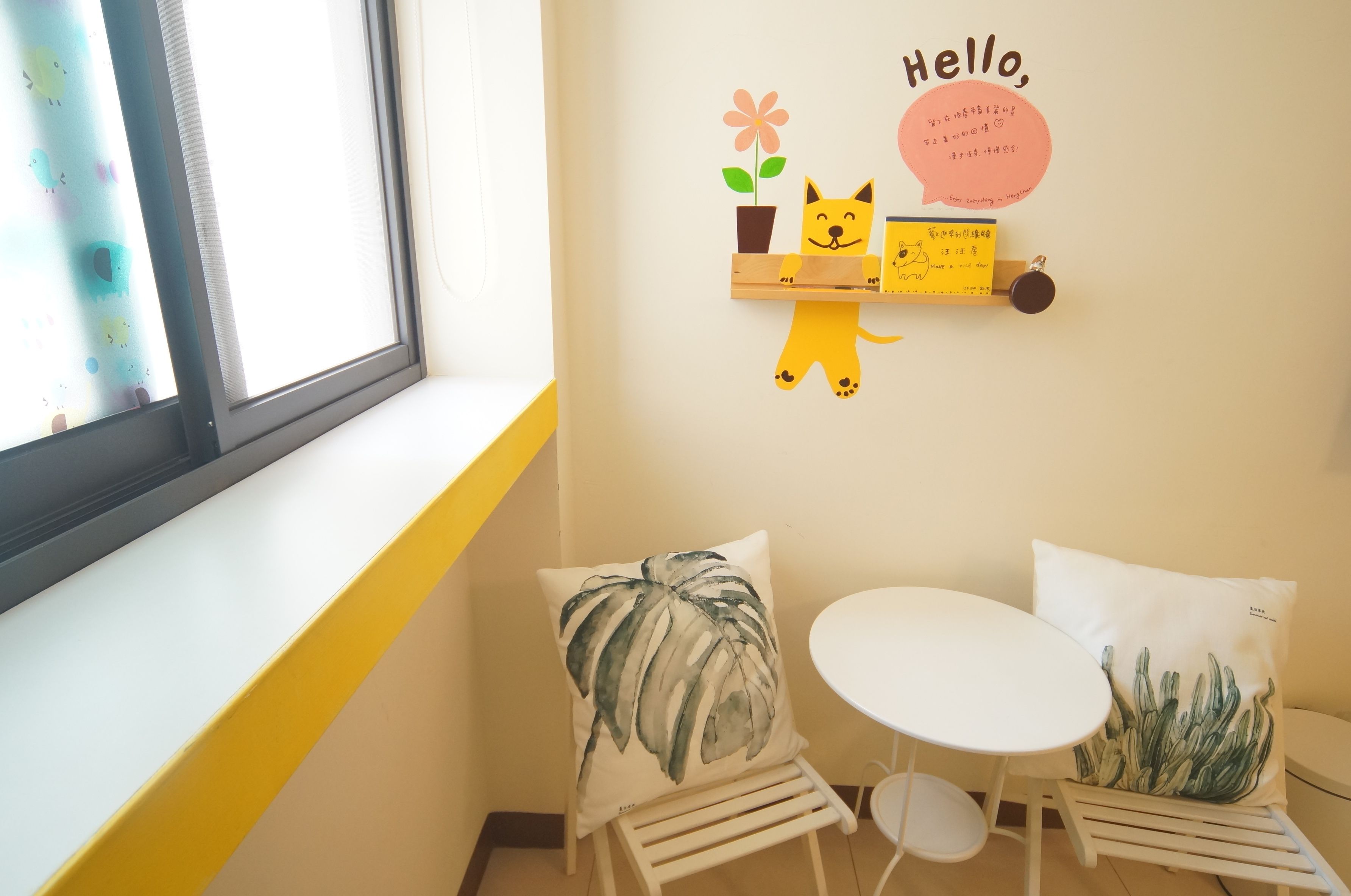 A small room with a window, a white chair, a small table, and decorative wall art including a cat, a flower, and a sign with Chinese characters.
