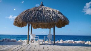 A wooden deck with a thatched roof structure, two white chairs, and a view of the ocean. - The Hill 25 – Elegant Apartment Near Blue Bay Beach & Golf Course, Curaçao (Sint Michiel)