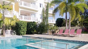 A swimming pool with a tiled deck, surrounded by palm trees and a multi-story building with balconies. - The Hill 25 – Elegant Apartment Near Blue Bay Beach & Golf Course, Curaçao (Sint Michiel)