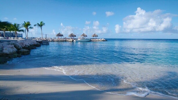 A beach with clear water, palm trees, and thatched-roof structures. - The Hill 25 – Elegant Apartment Near Blue Bay Beach & Golf Course, Curaçao (Sint Michiel)