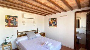 Un dormitorio con techo de madera, una cama con ropa blanca, dos lámparas fijadas a la pared y dos cuadros.