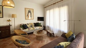 Una sala de estar con un sofá, dos sillas de mimbre, una mesa de centro, una lámpara colgante, un cuadro, un televisor y una ventana con cortinas.